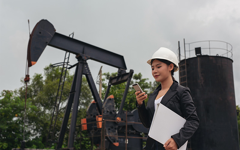 Person in safety helmet and formal attire holding phone and folder at oil extraction site with pump jacks.